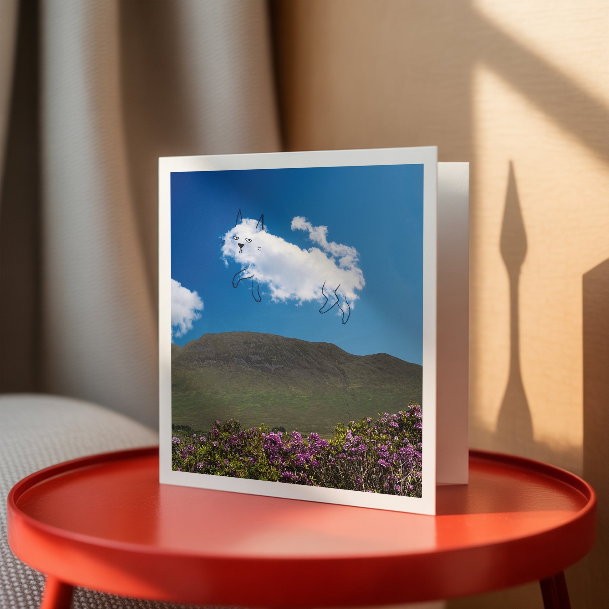 Greeting card with a cloud cat design on a scenic background, placed on a red table.