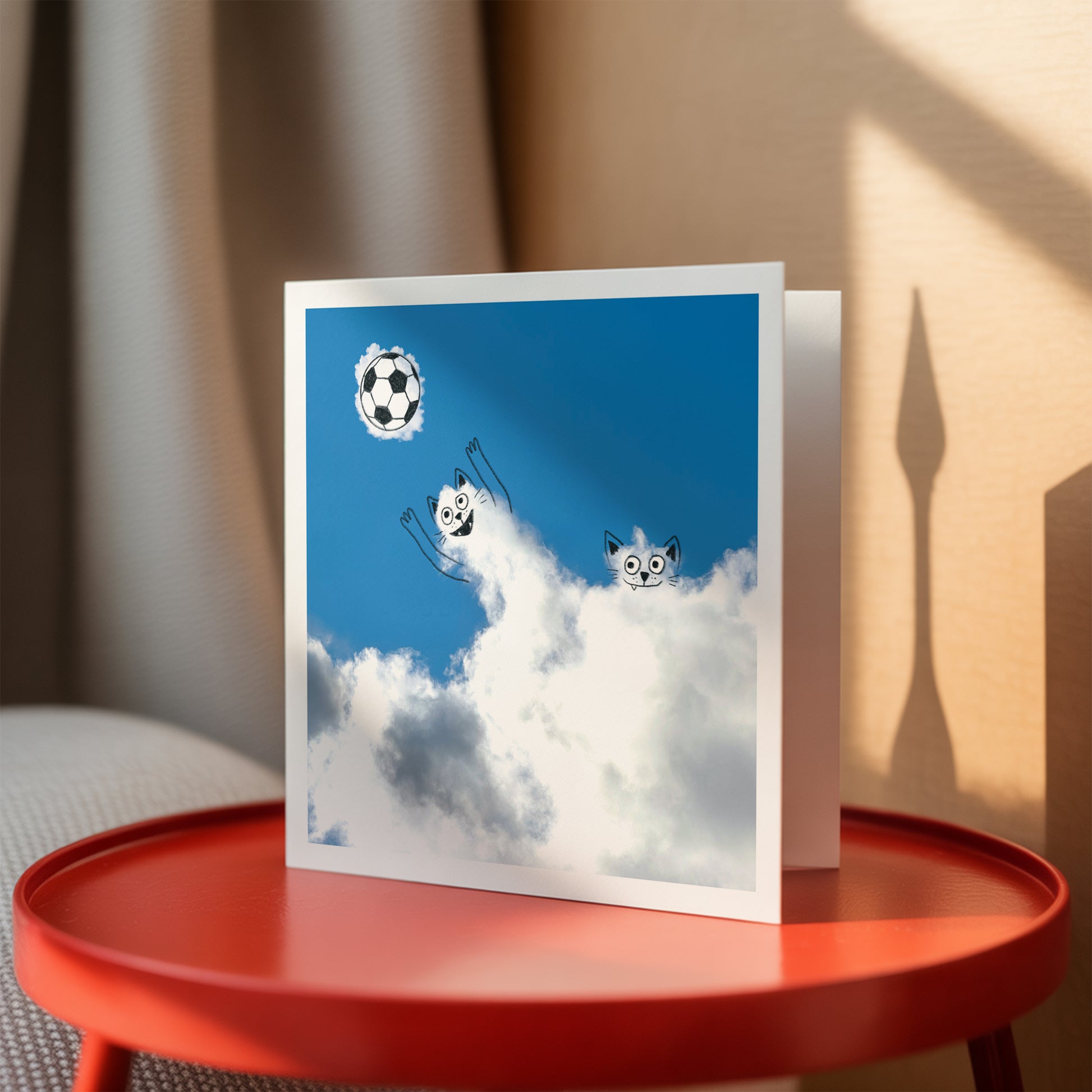 Greeting card with cats playing soccer in the sky on a red table.