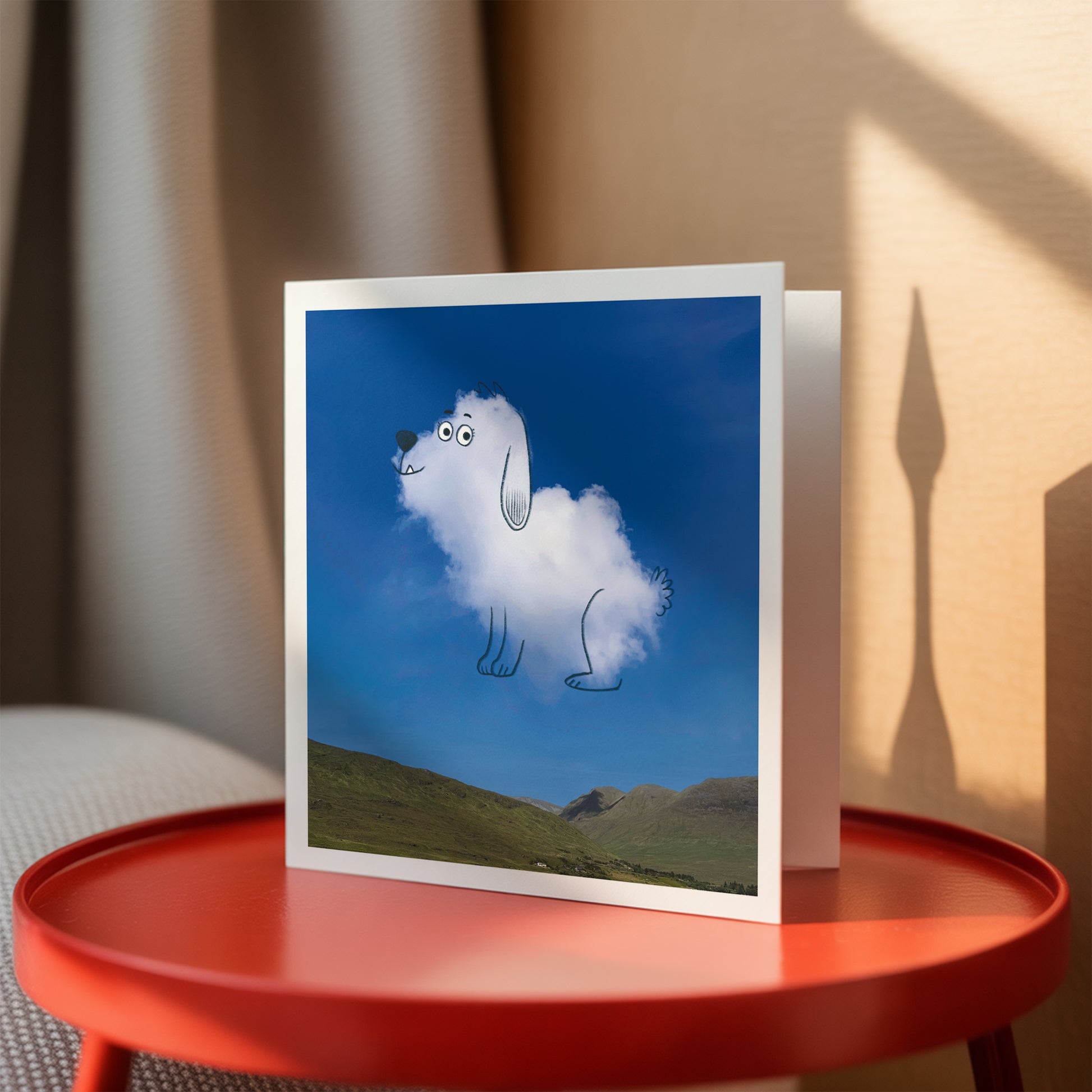 Greeting card with a cloud-shaped dog on a scenic background, placed on a red table.